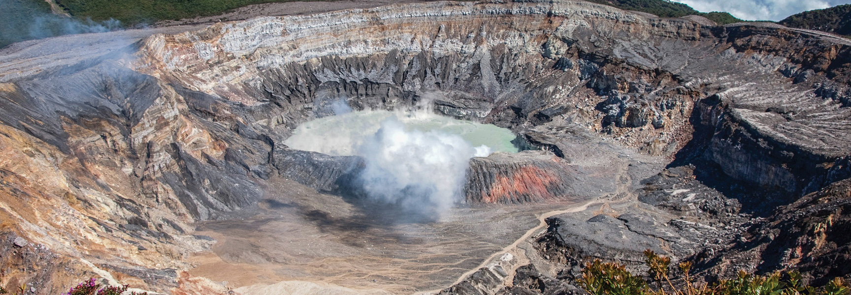 A wide-angle view of a steaming volcanic crater lake in Costa Rica with steep, multicolored rock walls.