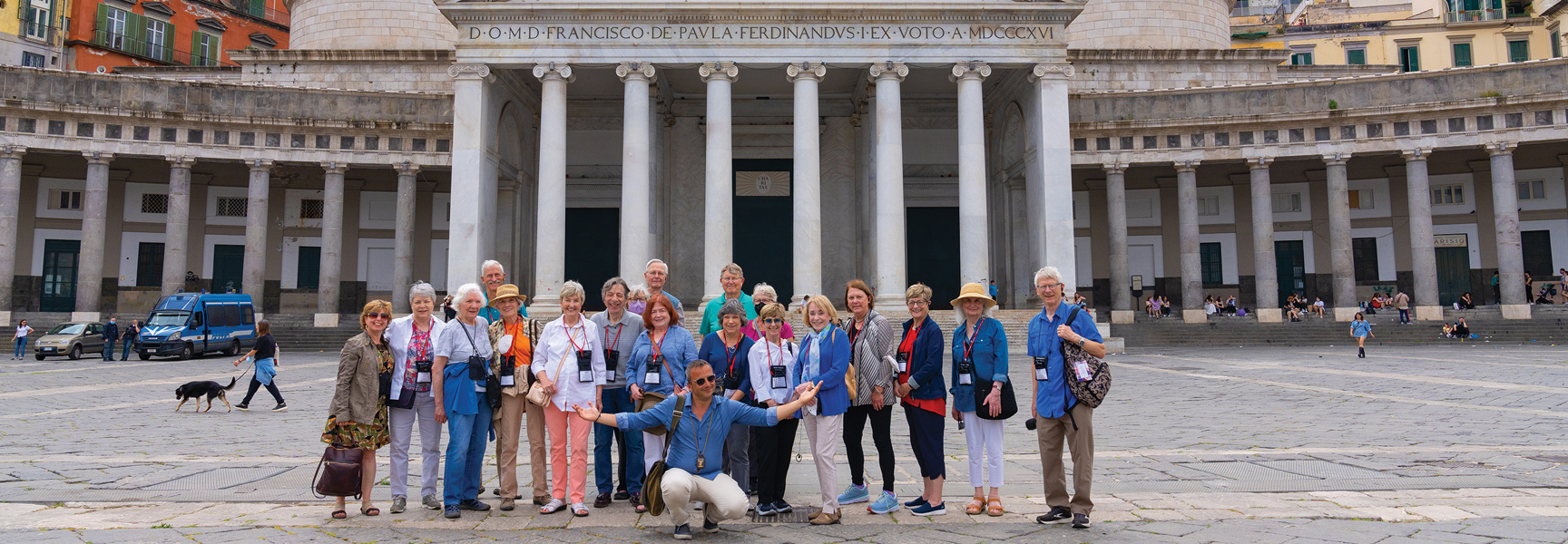 A group of travelers poses for a photo in a large piazza in front of a historic, colonnaded building in Naples, Italy.