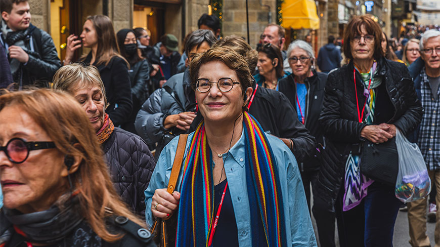 A smiling woman wearing glasses and a colorful scarf walks with a group through a crowded street in Florence.