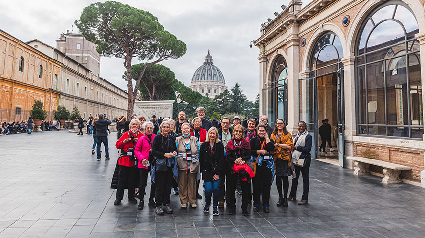 A group of people poses for a photo in a courtyard at The Vatican in Rome with St. Peter's Basilica visible in the background.