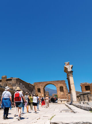 Tourists walk past tall ancient columns and stone ruins under a clear blue sky in Italy during a study program in Sorrento.