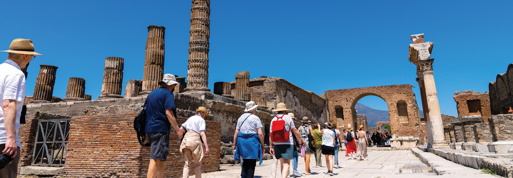 A group of people walks along a stone path through ancient ruins with large columns on a sunny day in Italy.