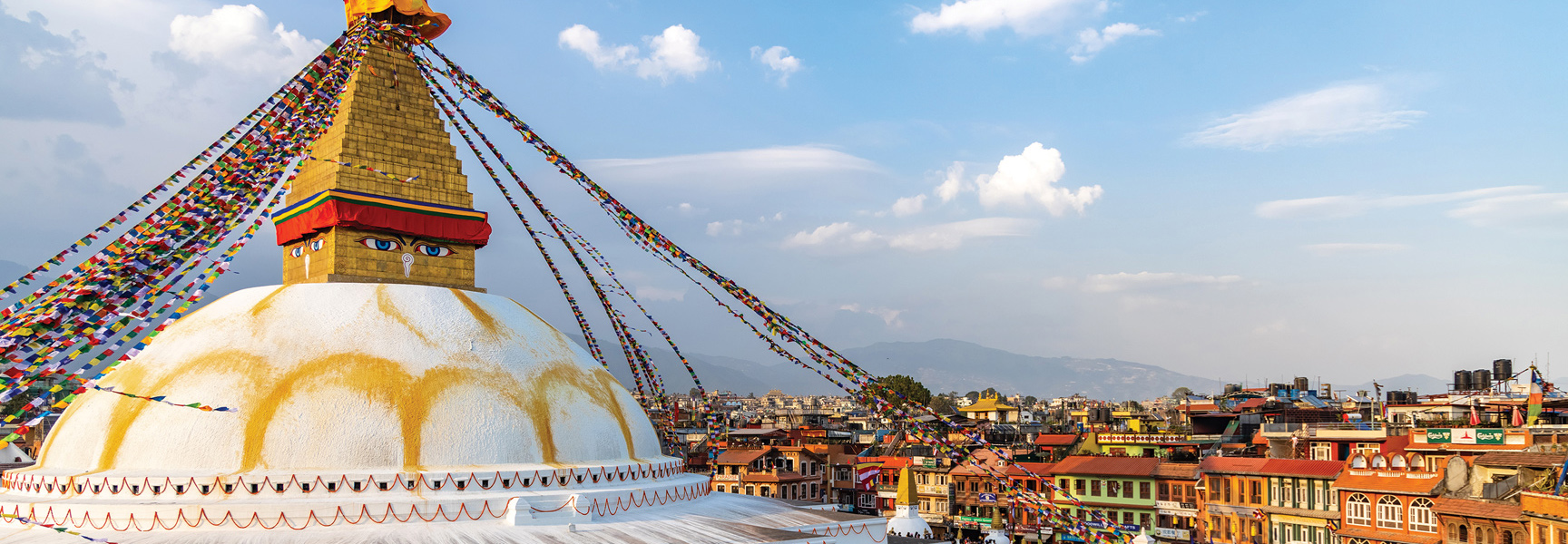 A large, white and gold stupa with colorful prayer flags overlooks the cityscape and distant mountains of Nepal.