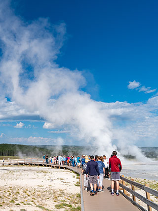 A group of tourists walks along a wooden boardwalk to view a steaming geyser erupting in Yellowstone National Park, Montana.