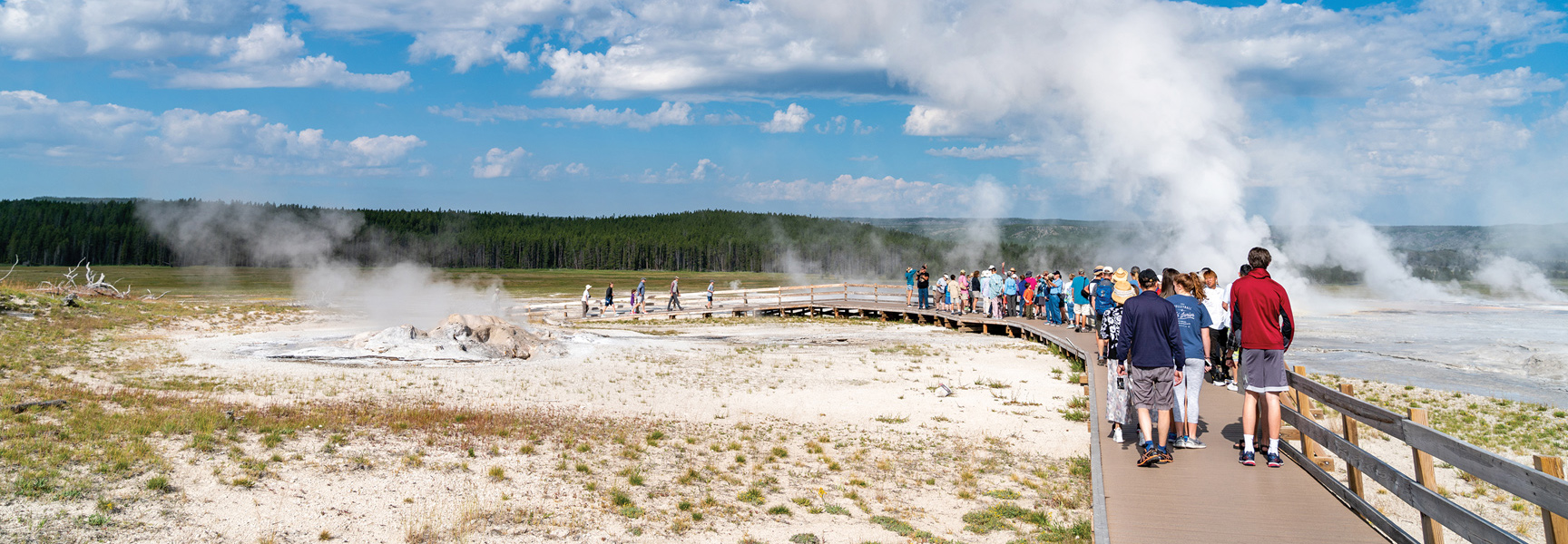 A crowd of tourists walks along a wooden boardwalk winding through a steaming geothermal basin in Yellowstone National Park, Montana.