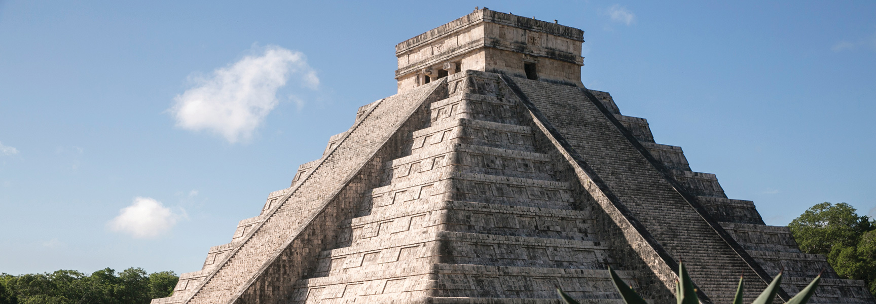 A large stone step-pyramid at a Mayan archaeological site in Mexico under a partly cloudy blue sky.
