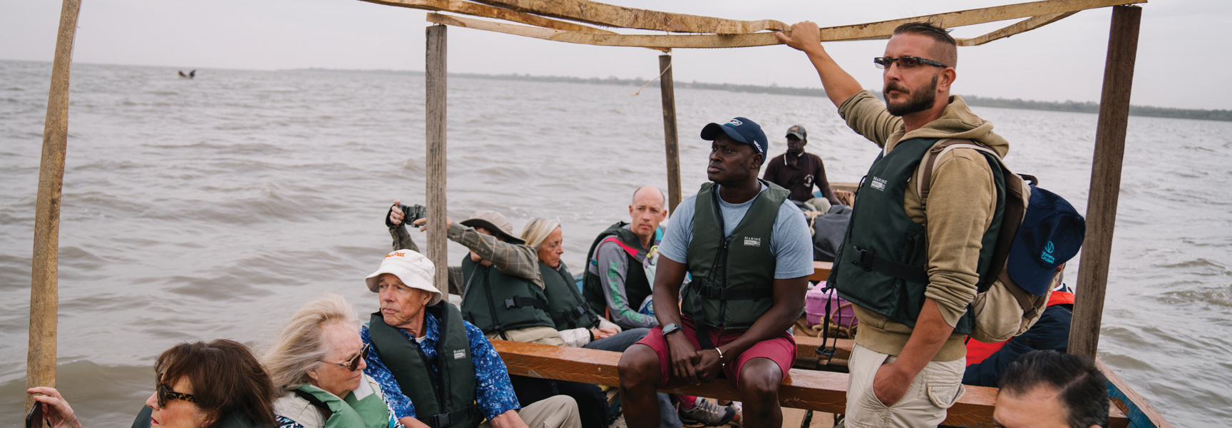 A group of travelers wearing life vests sit in a simple wooden boat on a river in Gambia/Senegal under an overcast sky.