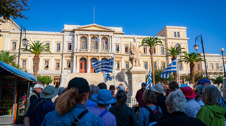 A group of travelers on a walking tour look up at the grand Ermoupoli Town Hall on the island of Syros, Greece.
