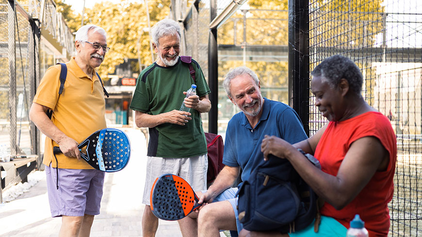 A group of four seniors in athletic wear laugh together by a pickleball court, some holding paddles and water.