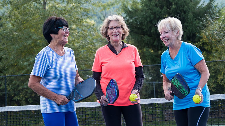 Three senior women laugh together on a pickleball court while holding their paddles and pickleballs.