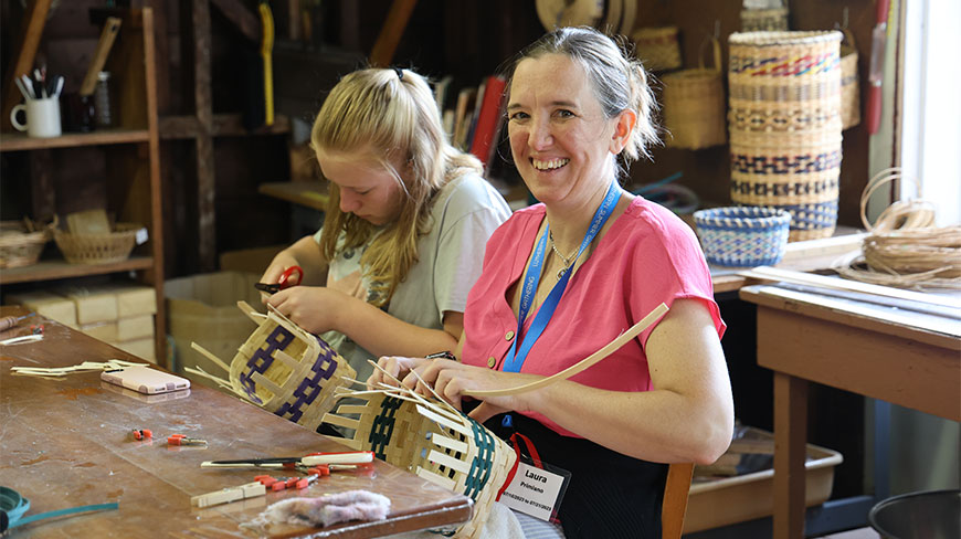 A smiling woman and a girl participate in a basket weaving workshop in New York, crafting handmade baskets at a wooden table.