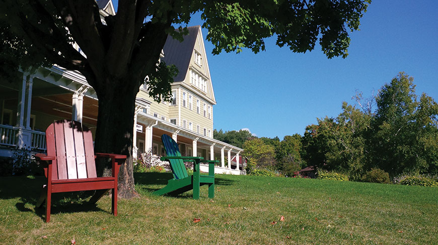 Two colorful Adirondack chairs sit on a green lawn under a shady tree in front of the Silver Bay YMCA Inn.