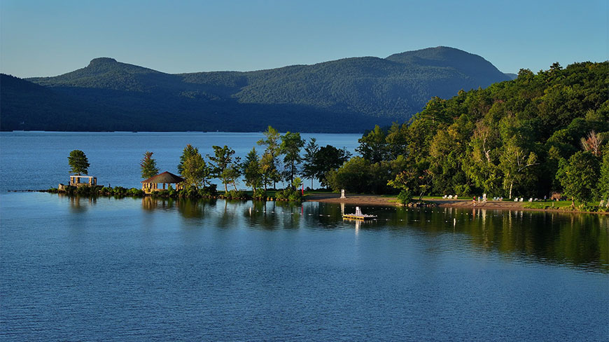 Slim Point Beach in New York features a gazebo on a peninsula and a calm swimming area surrounded by mountains and dense forests.