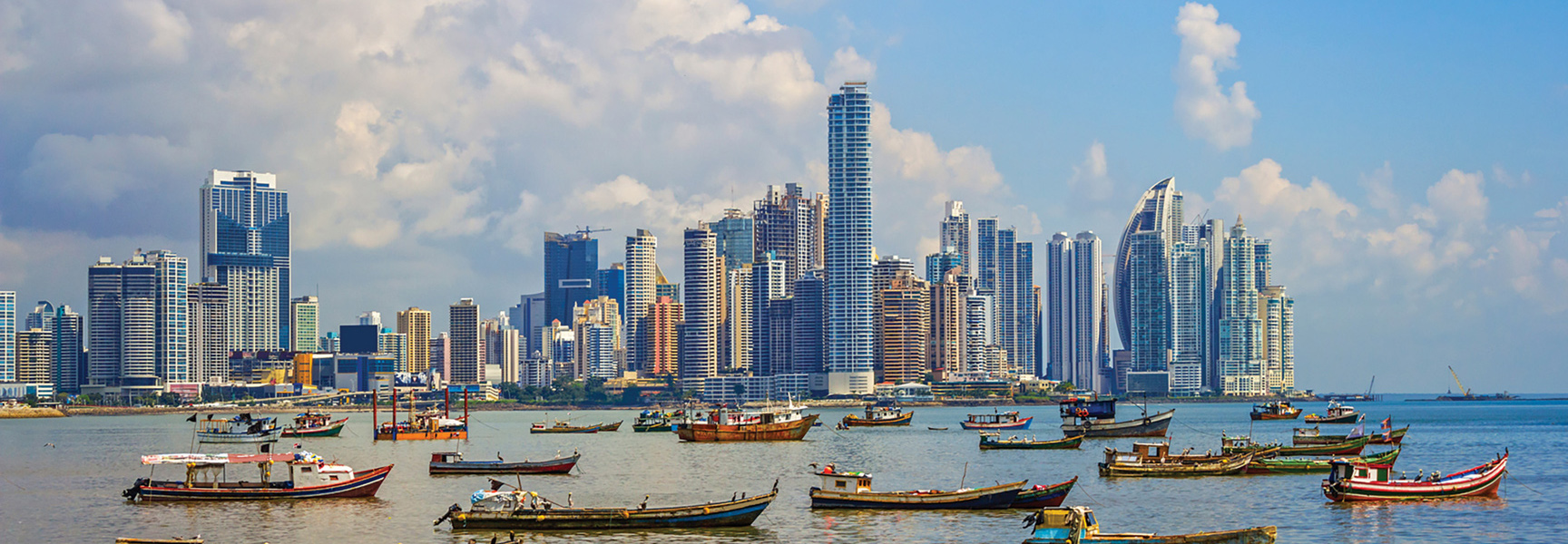 The modern skyline of Panama City, Panama, rises behind a harbor full of small fishing boats under a partly cloudy sky.