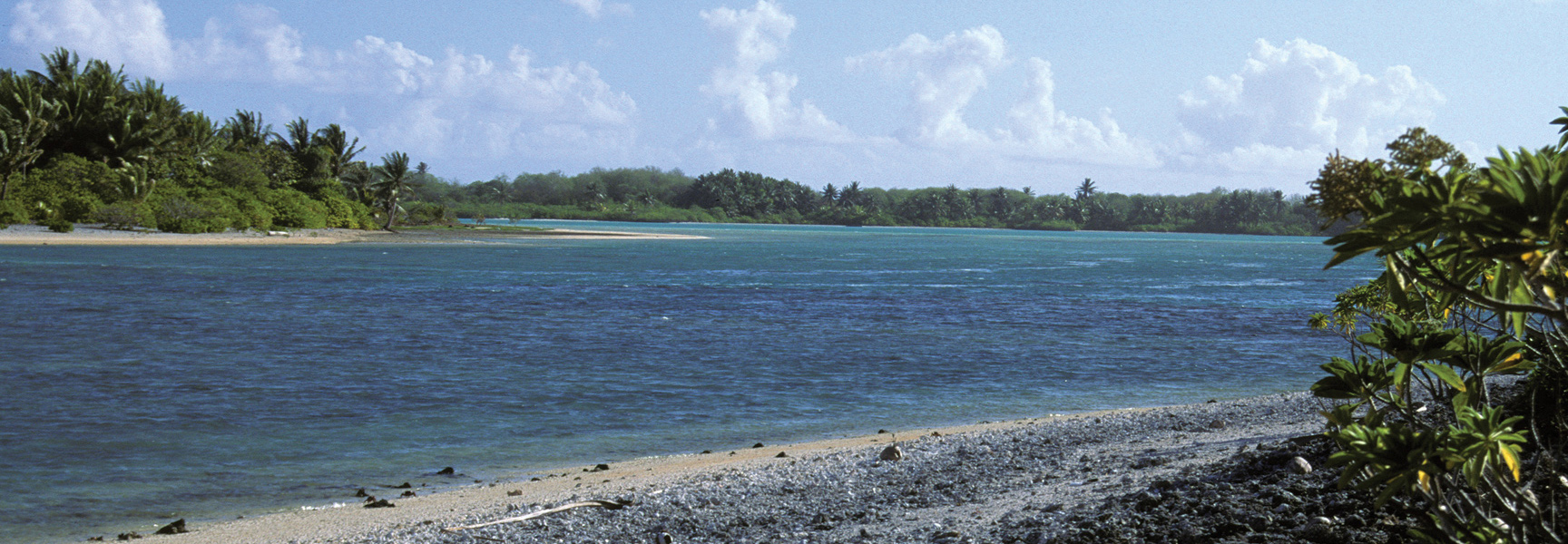 A gravel beach on a tropical island looks out over a calm turquoise lagoon toward a distant shore lined with palm trees.