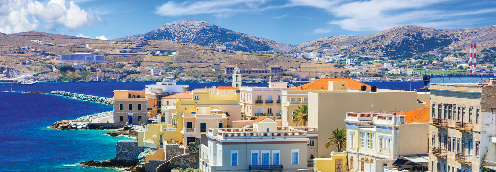 A colorful coastal town in Greece sits by the blue sea at the base of large, arid hills.