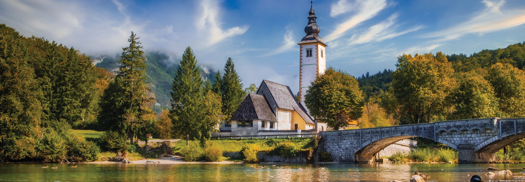 A picturesque church with a tall steeple and a stone bridge sits by a tranquil lake surrounded by lush, green forests and mountains.
