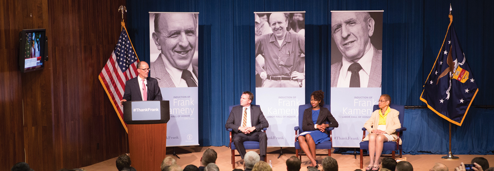 A speaker stands at a podium next to a seated panel on a stage, with large portraits of Frank Kameny in the background.