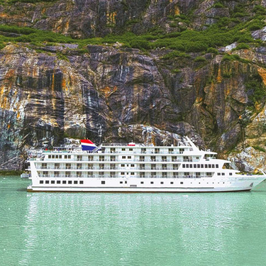 The American Constitution cruise ship sails on the green waters of an Alaskan fjord, dwarfed by the towering, colorful cliffs of the Inside Passage.