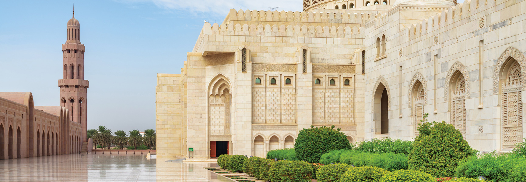 The ornate stone exterior of a large mosque with a minaret and manicured gardens on a clear day in Oman.