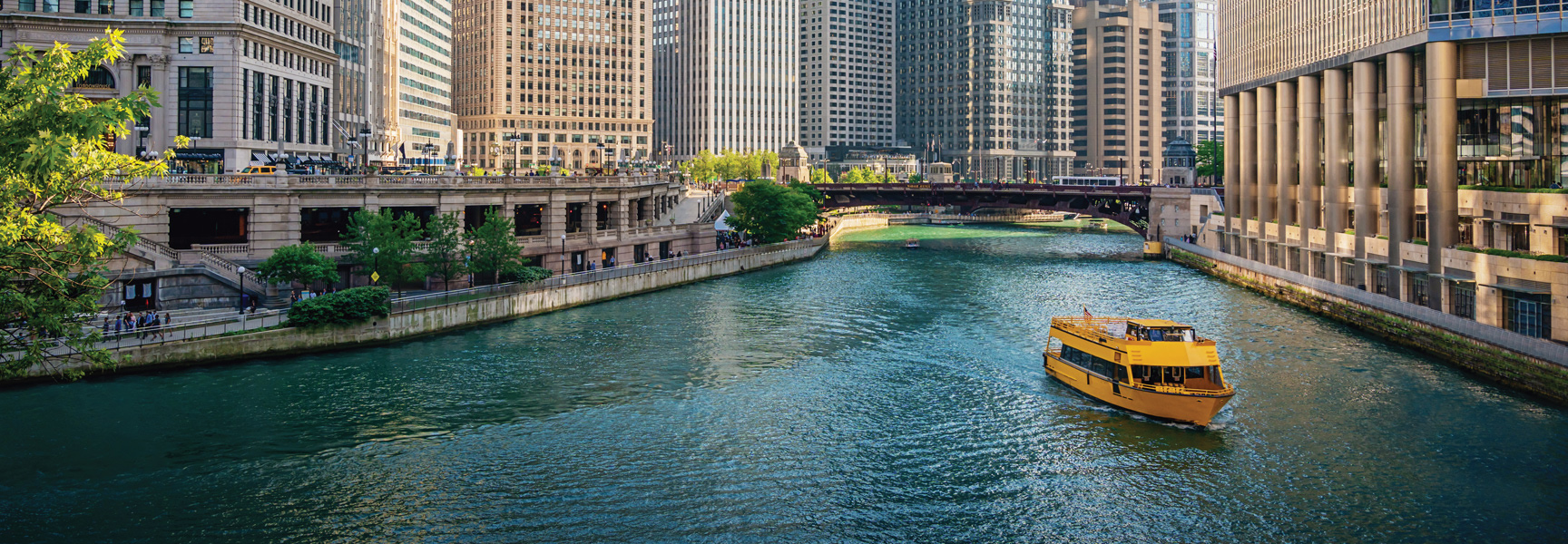 A yellow tour boat floats down the Chicago River, surrounded by the city's tall buildings and a bridge in the background.