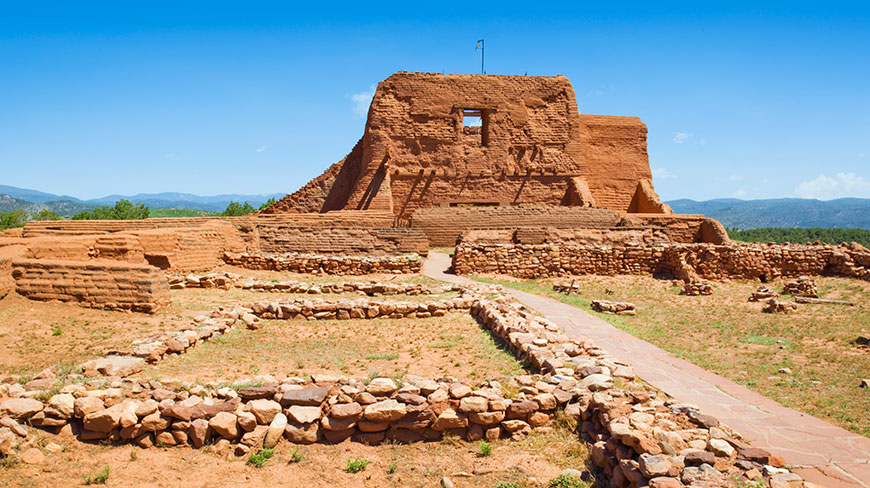 The red adobe ruins of the Pecos National Monument mission church and surrounding stone walls stand under a vast, clear blue sky.