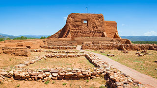 The reddish-brown adobe ruins of the Pecos National Monument mission church stand under a bright blue sky with low stone foundations in the foreground.