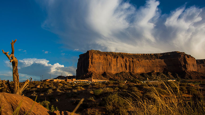 A large red rock mesa rises from the desert floor under a vast, cloud-streaked blue sky, with a dead tree to the left.