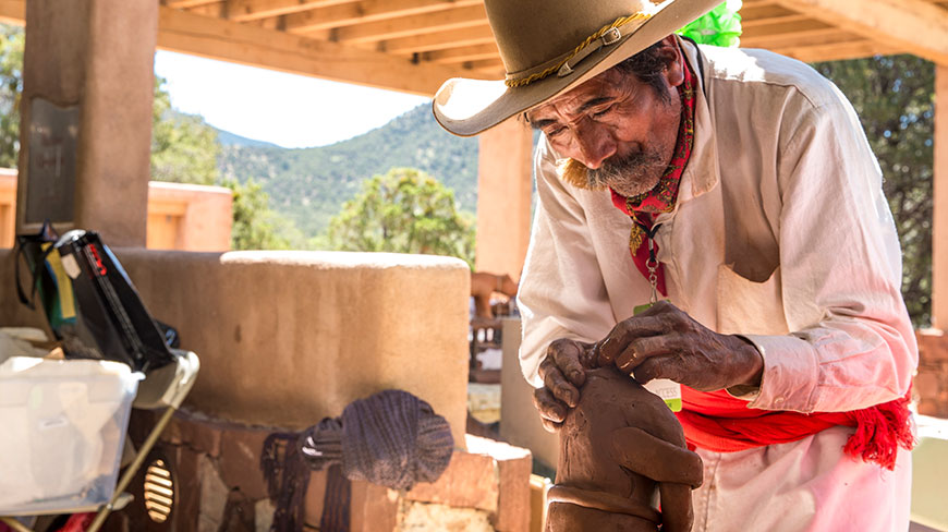 A man in a wide-brimmed hat carefully sculpts a piece of clay pottery outdoors in Santa Fe.