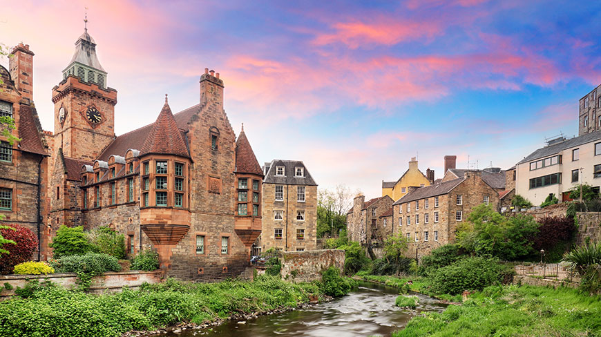 Historic stone buildings and a clock tower along the Water of Leith in Dean Village, Edinburgh, at sunset.