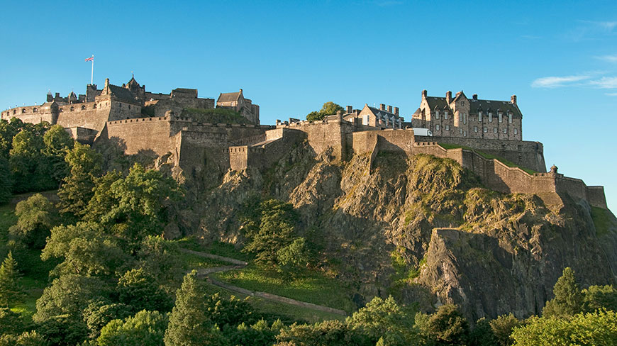 Edinburgh Castle sits atop a rocky cliff surrounded by lush green trees under a clear blue sky.