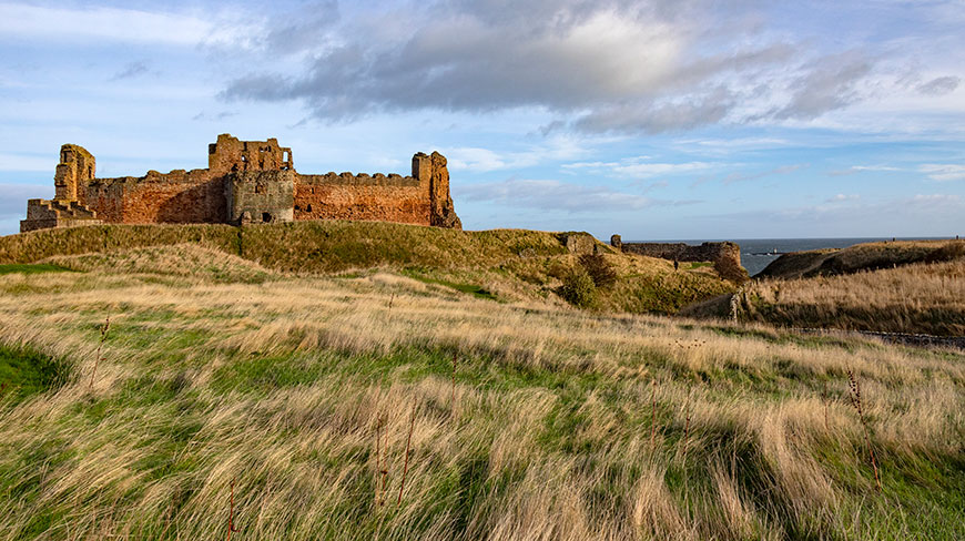 The ruins of Tantallon Castle sit atop a grassy hill overlooking the sea in Scotland under a partly cloudy sky.