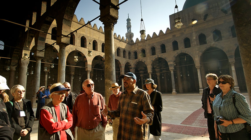 A tour guide gestures while speaking to a group of tourists in a stone courtyard of a historic building.