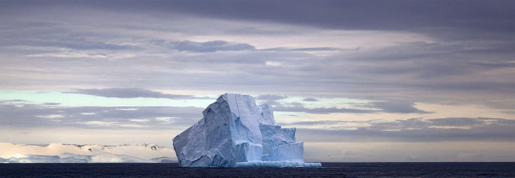 A large, craggy iceberg floats in the dark ocean water under a cloudy sky, with snow-covered land in the distance.
