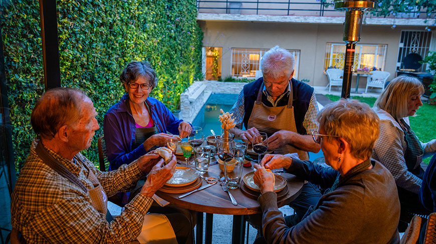 A group of seniors wearing aprons sit around a wooden table, enjoying an outdoor meal together on a patio at dusk.
