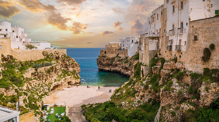 The coastal town of Polignano a Mare overlooks a small beach tucked between dramatic cliffs under a warm, cloudy sky.