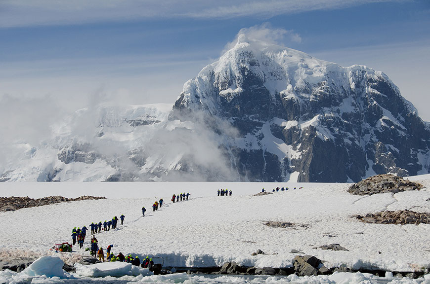 A group of explorers hike up a snowy Antarctic peninsula with a towering, cloud-wreathed mountain in the background under a blue sky.