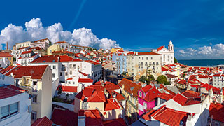 The dense, colorful buildings and red rooftops of Lisbon's Alfama district are pictured under a bright blue sky with puffy white clouds.