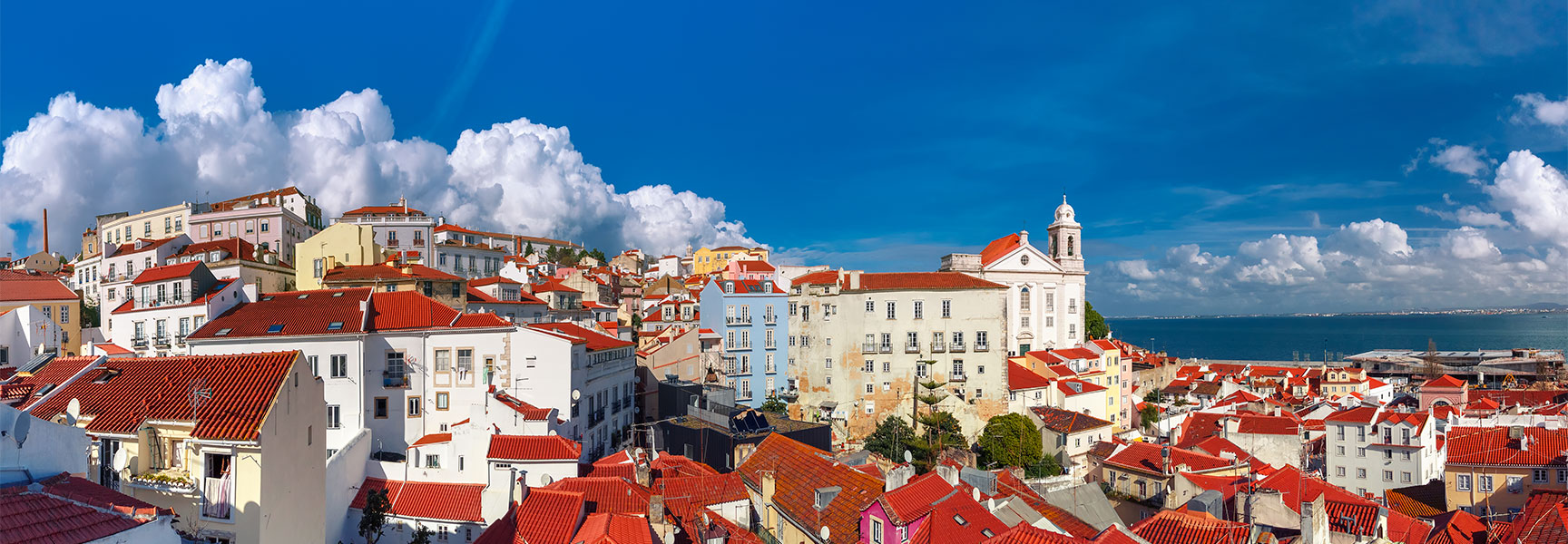 A panoramic view of Lisbon's Alfama district shows red-roofed buildings on a hill overlooking the ocean under a bright blue, cloud-filled sky.