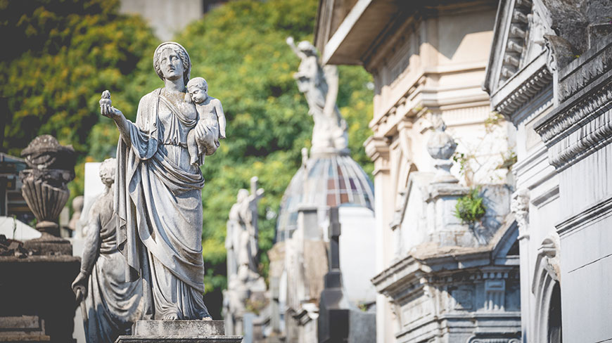A classical stone statue of a woman holding a child overlooks other elaborate mausoleums at the Recoleta Cemetery.