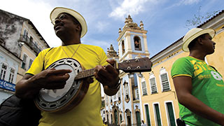 A man wearing a yellow shirt and a white fedora plays a banjo on a cobblestone street with colonial architecture in the background.