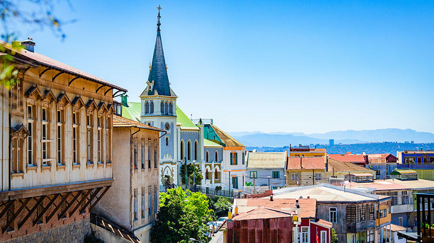A colorful Valparaiso hillside is dotted with historic buildings and a church steeple under a clear blue sky.