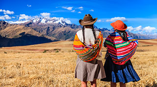 Two Peruvian women wearing traditional shawls and hats look out over a golden field toward a majestic, snow-capped mountain range under a clear blue sky.