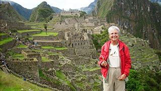 A woman in a red jacket smiles while standing in front of the ancient ruins and green terraced hills of Machu Picchu.