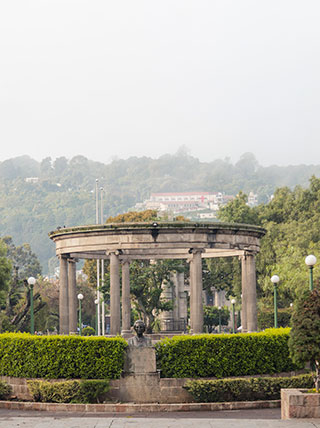 A circular stone kiosk with columns stands in a park with green hedges, with a forested hill in the background.