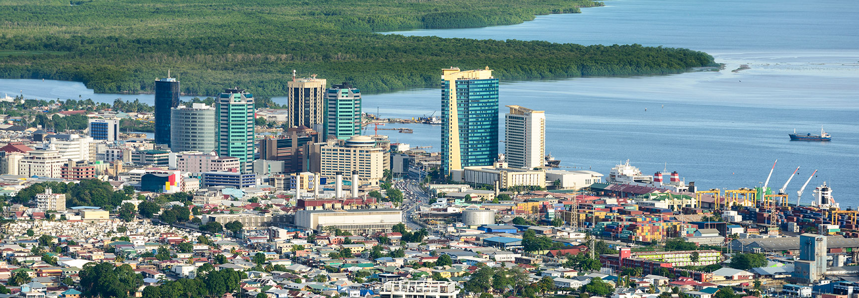 An aerial view of downtown Port of Spain, with modern skyscrapers along the waterfront next to a busy commercial port and lush green hills.