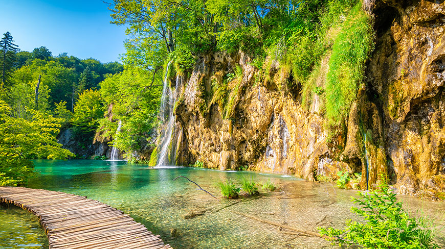 Waterfalls cascade down a lush, green cliff into a clear turquoise lake with a wooden boardwalk in the foreground.