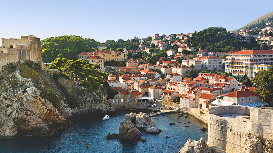 A scenic view of the old city of Dubrovnik, Croatia, with its stone walls and red-tiled roofs overlooking a calm blue bay.