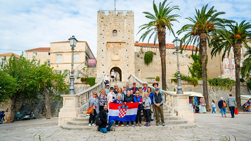 A tour group holds up a Croatian flag while posing for a photo on stone steps in front of a historic tower in Korcula.