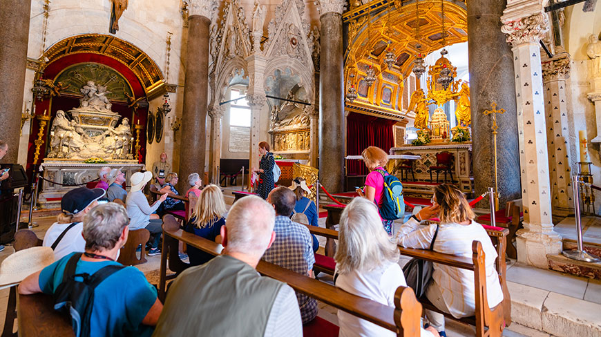 A tour group sits on wooden pews listening to a guide inside the ornate, historic interior of the Cathedral of Saint Domnius in Split.